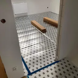 Przygotowanie do układania paneli podłogowych: folia izolacyjna rozłożona na podłodze, deski paneli ułożone w stosy, widok z perspektywy wejścia do pokoju z widocznym grzejnikiem i oknami dachowymi.