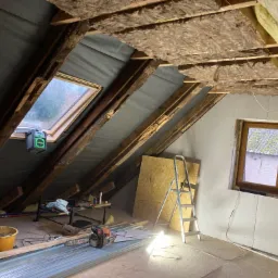 Poddasze w trakcie izolacji: widoczne wełna mineralna między krokwiami, folia paroizolacyjna, okno dachowe z laserowym niwelatorem, drabina, płyty OSB oparte o ścianę, narzędzia i elementy...
