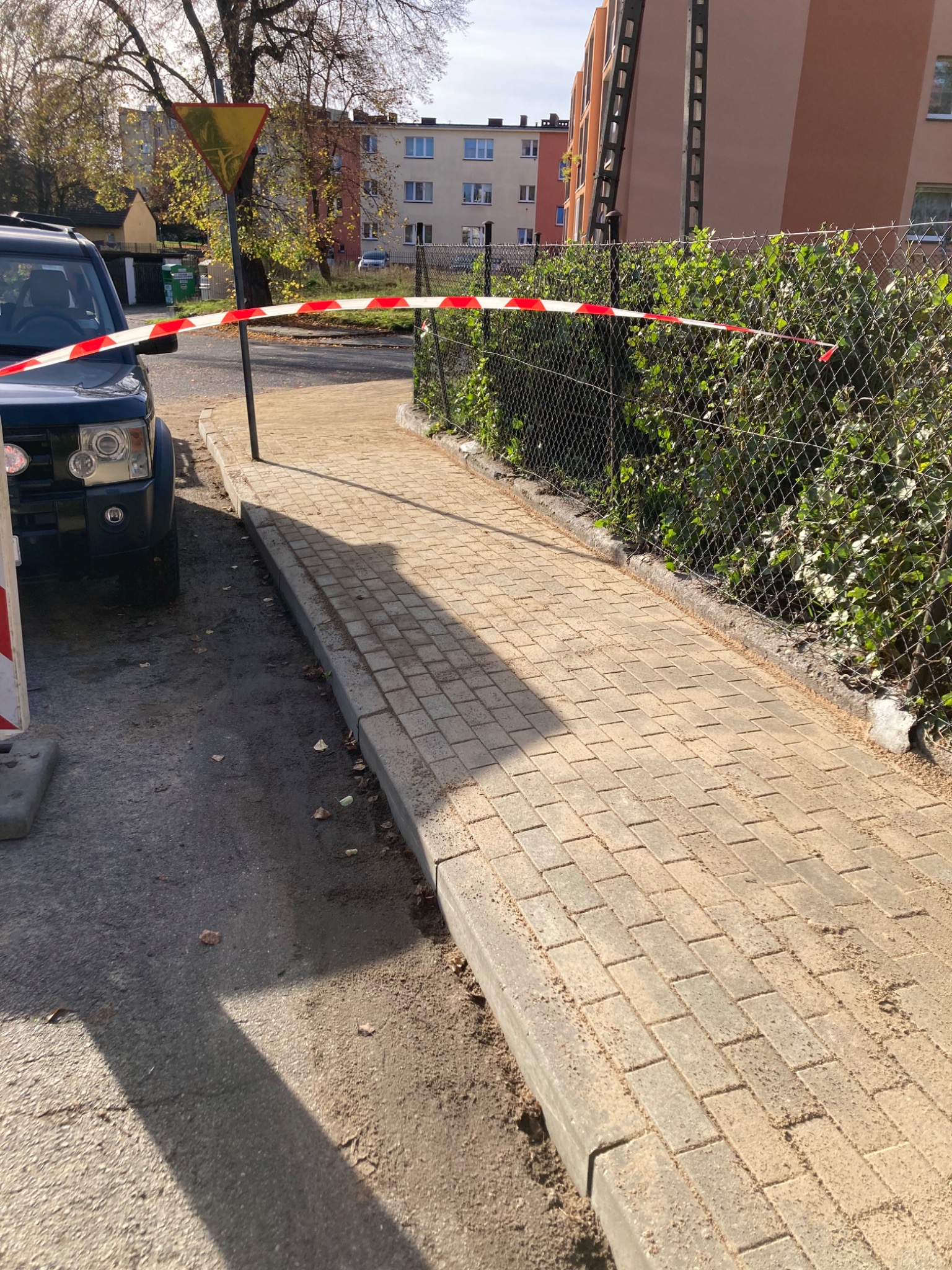 Nowo ułożony chodnik z kostki brukowej, odgrodzony taśmą ostrzegawczą, z widocznym znakiem drogowym 'ustąp pierwszeństwa' i fragmentem ciemnego samochodu terenowego po lewej stronie, na tle bloku...