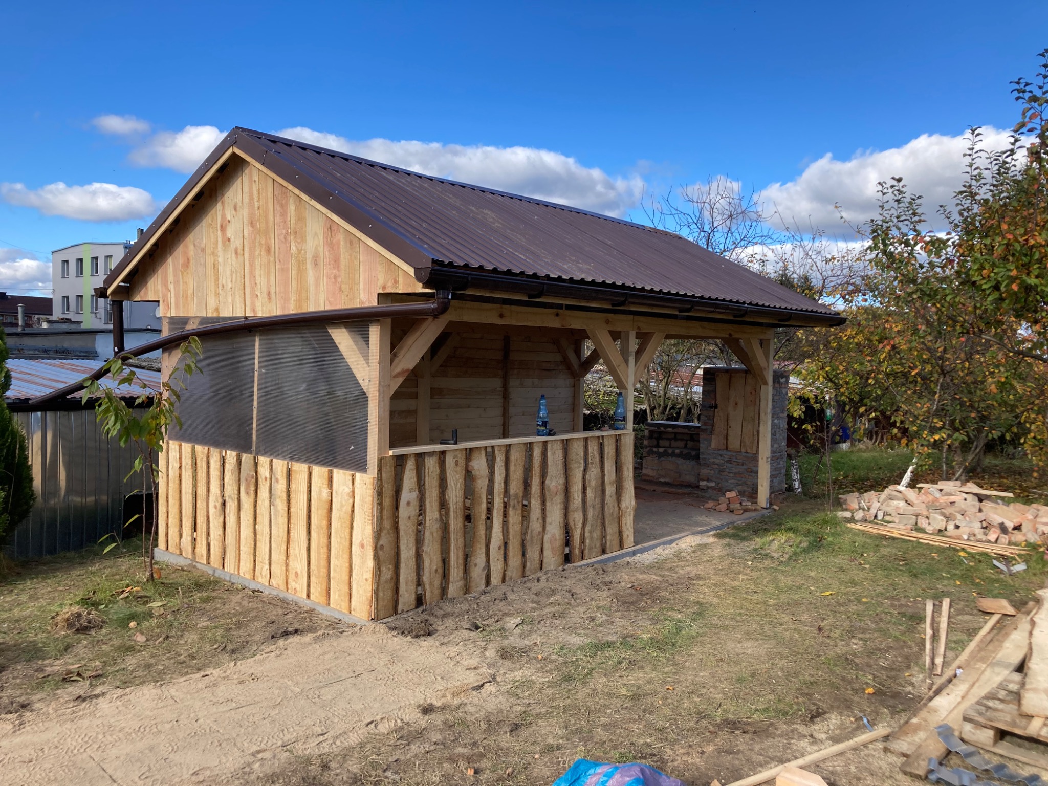 Drewniana altana z brązowym, metalowym dachem w trakcie budowy, widoczne surowe deski, częściowo osłonięta przezroczystym tworzywem, z paleniskiem w tle i stertą cegieł obok.