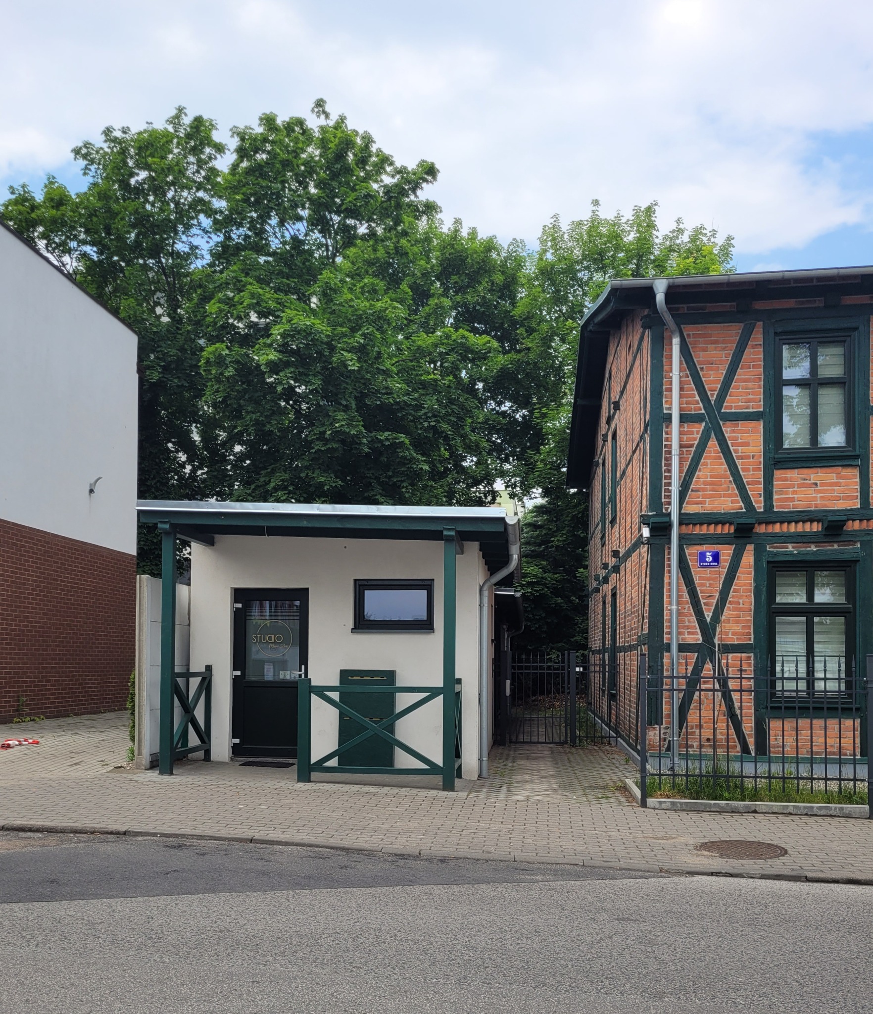 Zewnętrzny widok małego salonu kosmetycznego z zielonymi akcentami i napisem 'STUDIO' na drzwiach, usytuowanego obok budynku z czerwonej cegły z elementami szachulcowymi.