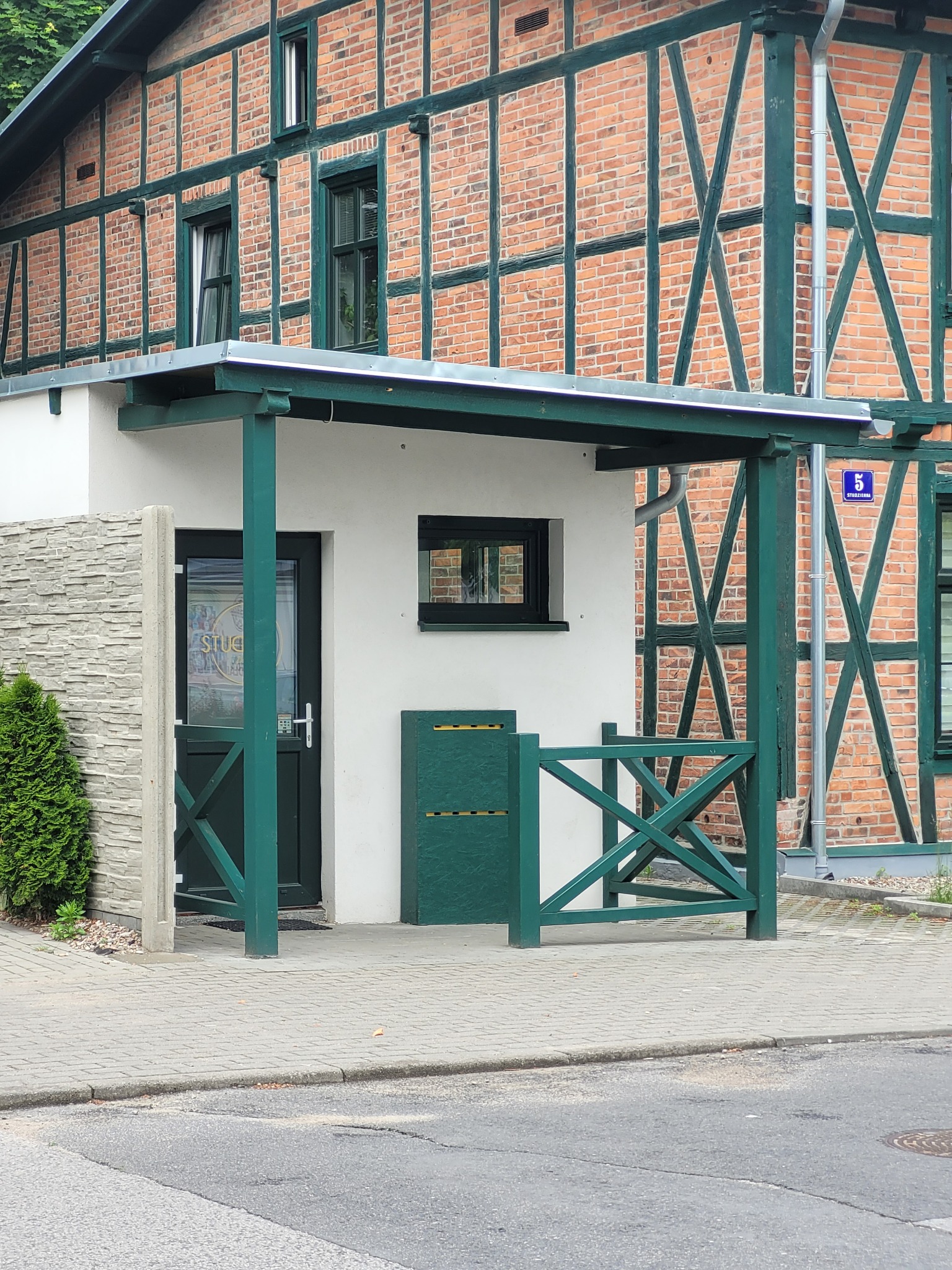 Wejście do budynku z zielonymi elementami konstrukcyjnymi i szyldem 'Studio' na szklanych drzwiach.
