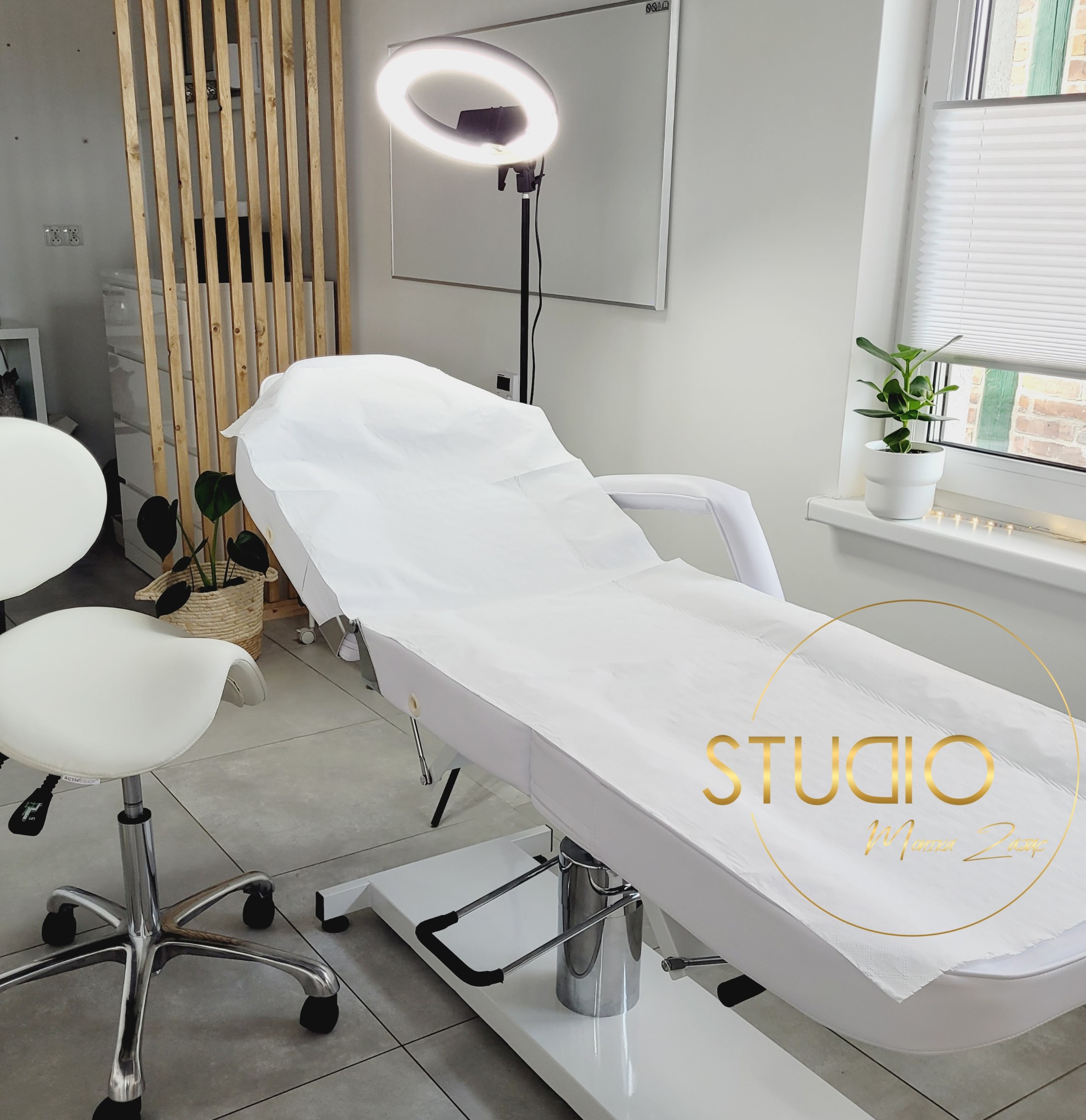 Białe krzesło i leżanka kosmetyczna z jednorazowym prześcieradłem w jasnym pomieszczeniu, okrągła lampa doświetlająca, roślina w doniczce na parapecie.