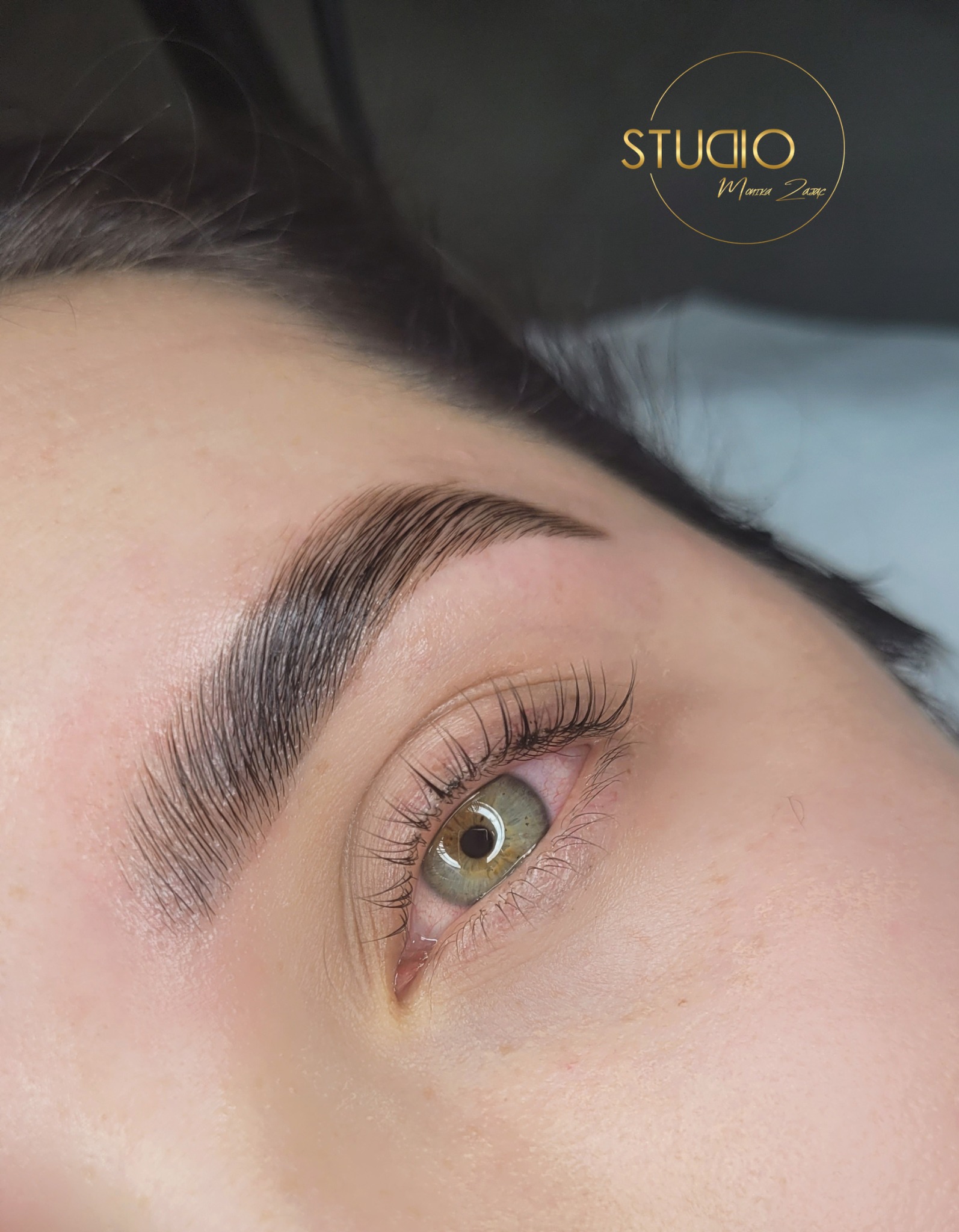 Zbliżenie na oko z widocznymi, wystylizowanymi brwiami po zabiegu laminacji, z delikatnym logo salonu w górnym rogu.