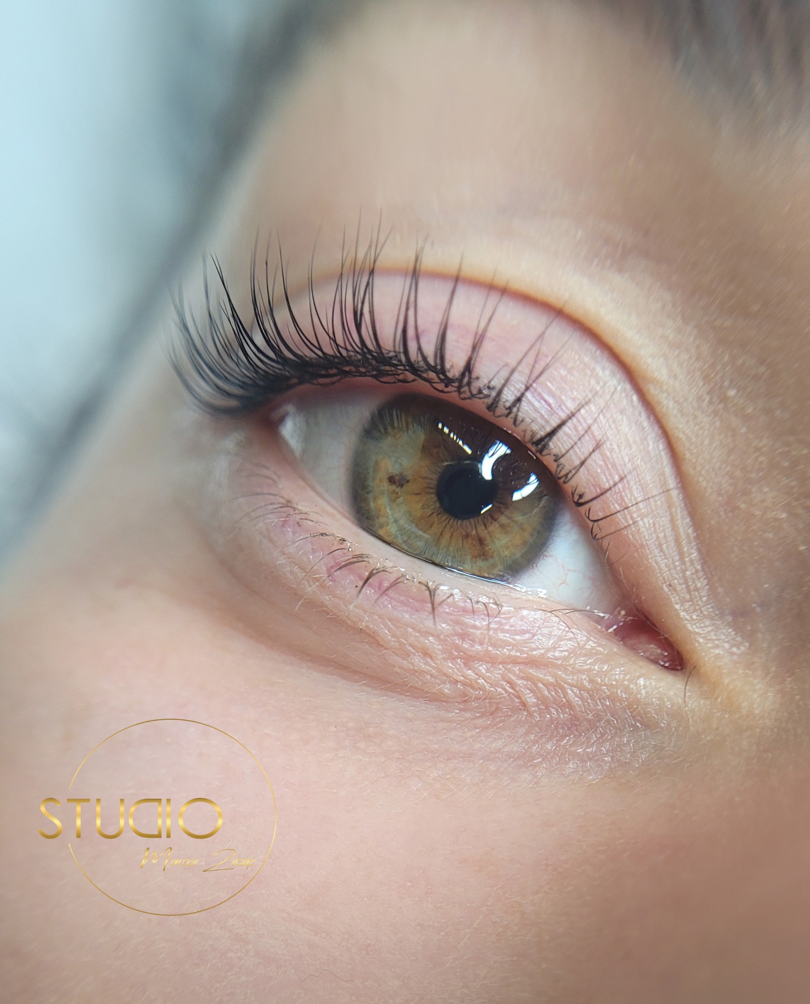 Zbliżenie na oko z brązową tęczówką i wydłużonymi, czarnymi rzęsami po zabiegu liftingu, widoczne logo studia w lewym dolnym rogu.