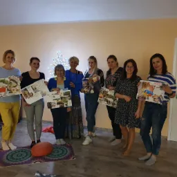 Grupa kobiet prezentuje kolaże z inspiracjami podczas warsztatów w jasnym pomieszczeniu z podświetlonym drzewkiem dekoracyjnym w tle.