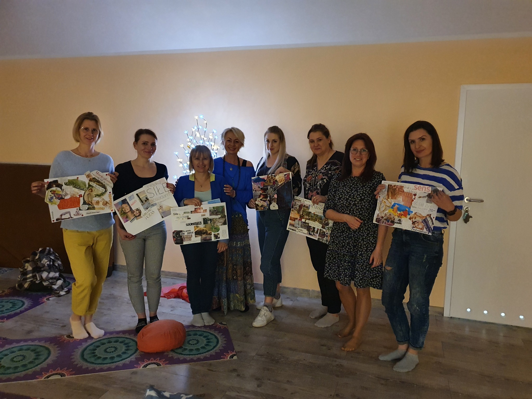 Grupa kobiet prezentuje kolaże z inspiracjami podczas warsztatów w jasnym pomieszczeniu z podświetlonym drzewkiem dekoracyjnym w tle.