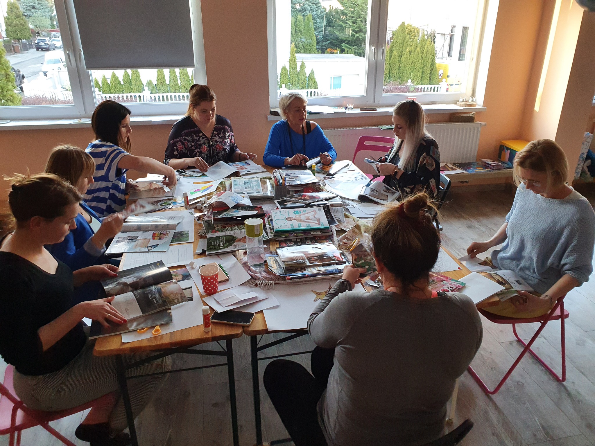 Grupa osób siedzi przy stole, wycinając i sklejając elementy z magazynów, tworząc kolaże w jasnym pomieszczeniu z dużymi oknami.