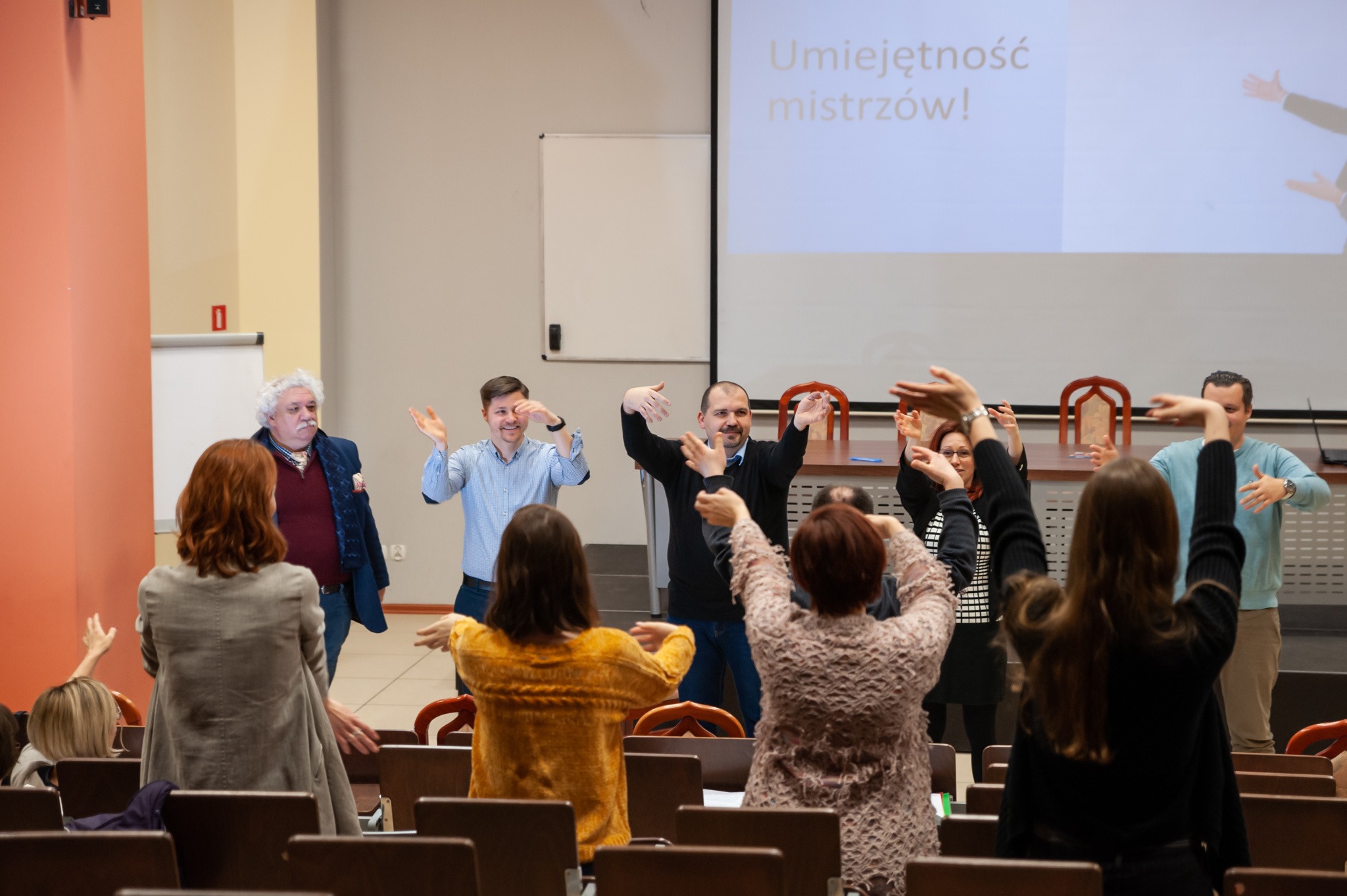Uczestnicy szkolenia w sali konferencyjnej wykonują ćwiczenie z podniesionymi rękami, na ekranie wyświetlony napis 'Umiejętność mistrzów!'