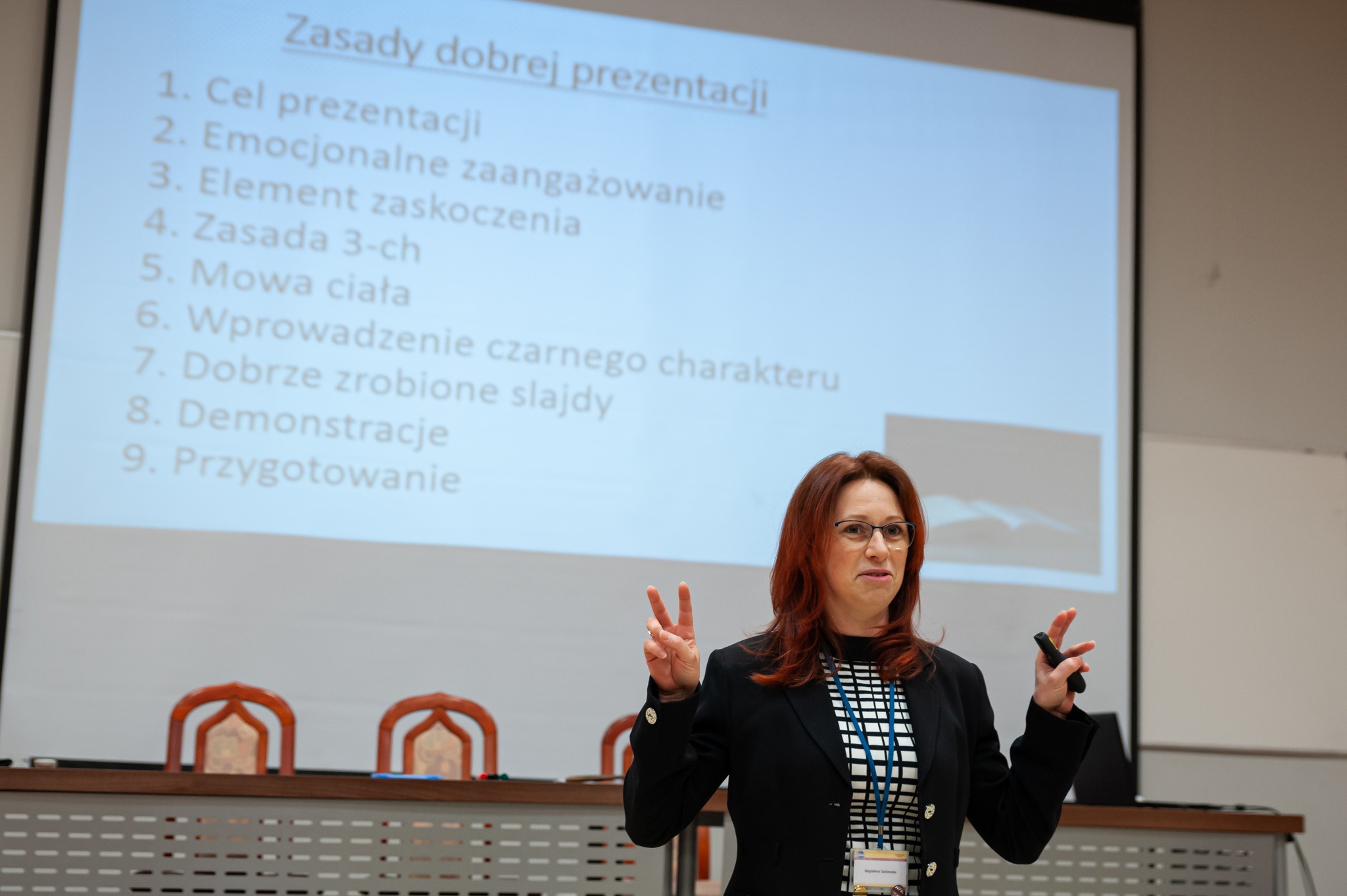 Prelegentka z czerwonymi włosami w okularach gestykuluje podczas prezentacji o zasadach dobrej prezentacji, wyświetlonych na ekranie za nią.