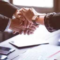 Złożone dłonie na biurku w Tuszynie symbolizujące współpracę, obok leżą smartfony, tablet, klawiatura i filiżanka kawy.