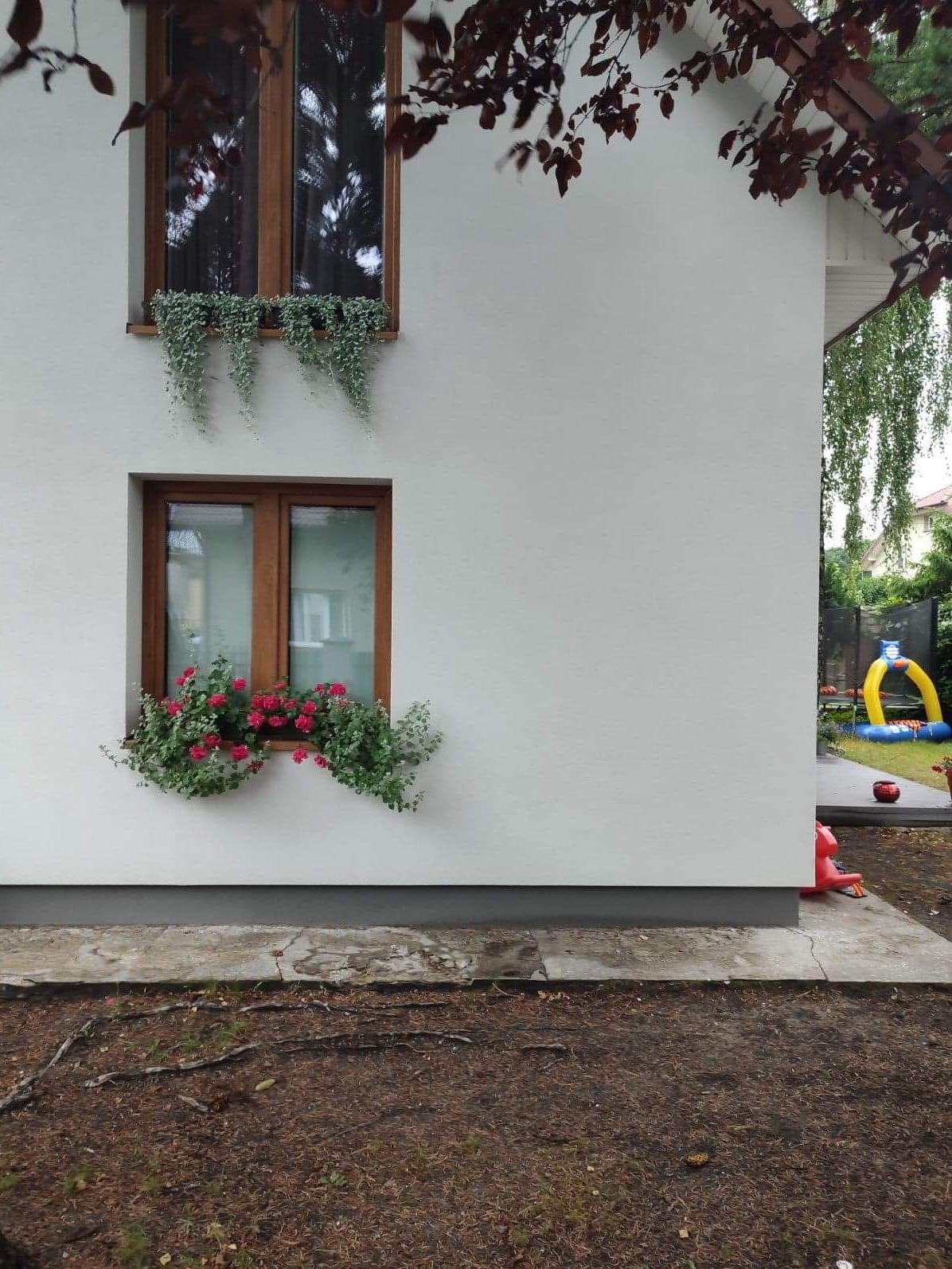 Boczny widok ocieplonego domu z białym tynkiem silikonowym, drewnianą stolarką okienną i kwiatami w donicach na parapecie, w tle widoczny plac zabaw.