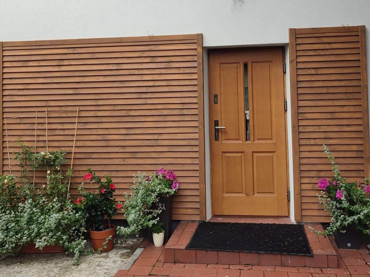 Wejście do domu z drewnianą elewacją z poziomych desek, brązowe drzwi, schody z czerwonej cegły, kwiaty w doniczkach po obu stronach wejścia.
