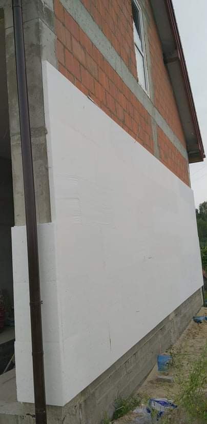 Fragment ściany budynku z czerwonej cegły z nałożonymi białymi płytami izolacyjnymi, widoczna rynna i okno na górze.