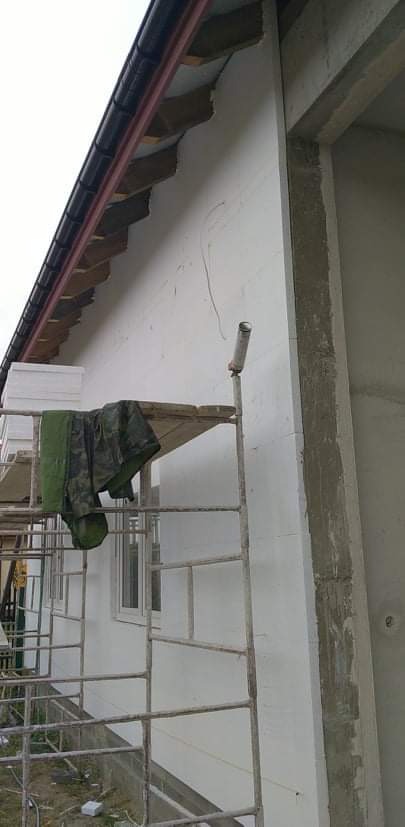 Ściana budynku w trakcie ocieplania z widocznymi rusztowaniami, narzędziami i fragmentem elewacji z zamontowanym oknem.