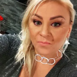 Portret kobiety z blond włosami, subtelnym makijażem oczu w odcieniach różu i srebrną biżuterią, w błyszczącej, ciemnej kreacji, na tle ściany imitującej kamień.