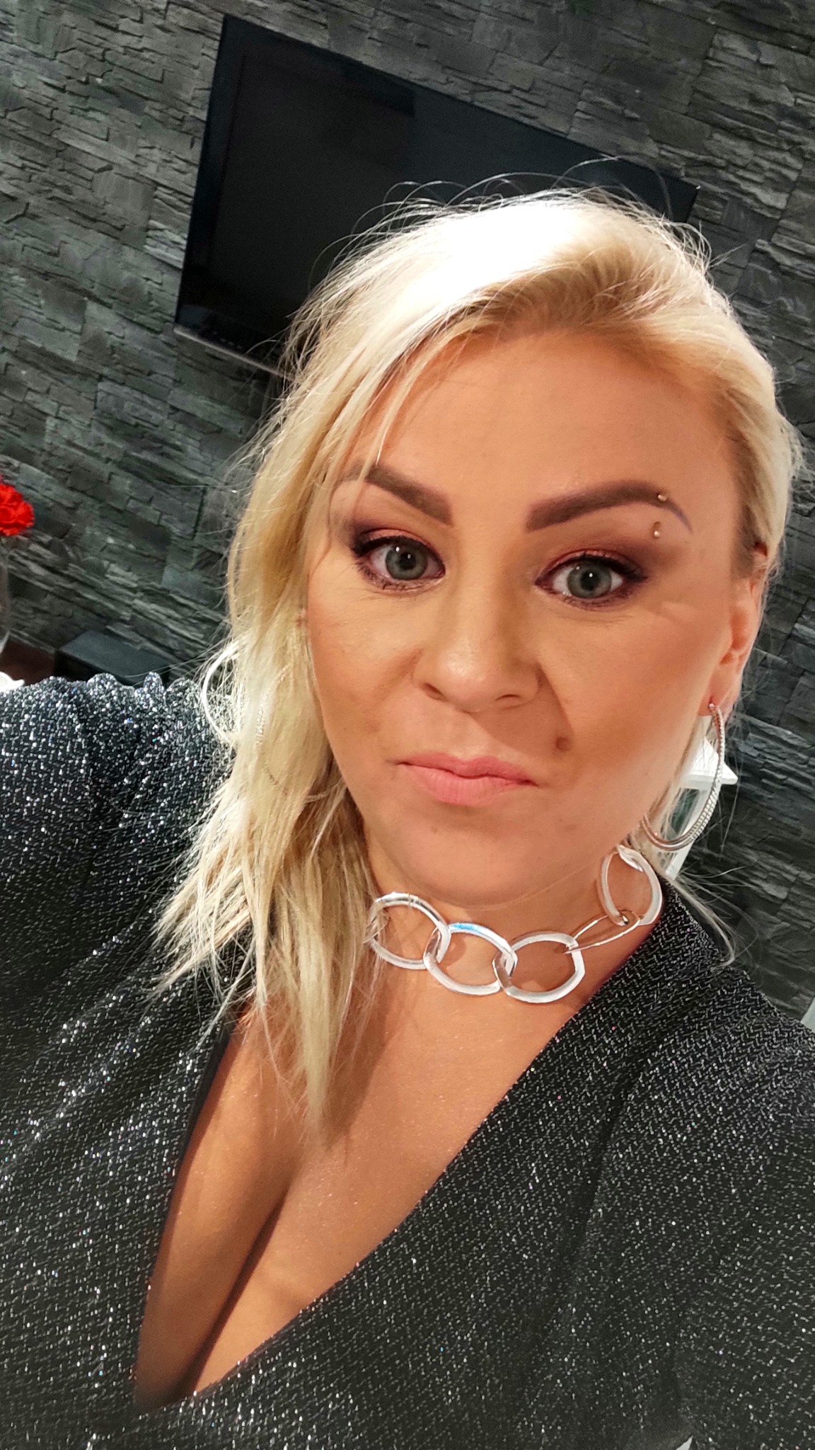 Portret kobiety z blond włosami, subtelnym makijażem oczu w odcieniach różu i srebrną biżuterią, w błyszczącej, ciemnej kreacji, na tle ściany imitującej kamień.