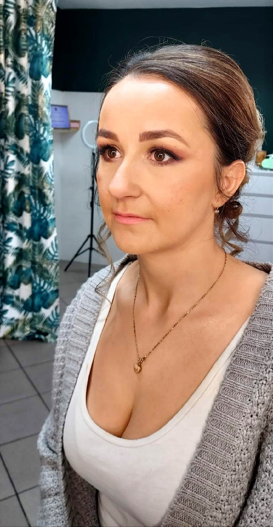 Kobieta z delikatnym makijażem, widoczny róż na powiekach i podkreślone kości policzkowe, ubrana w szary sweter i biały top, z złotym naszyjnikiem, w tle zasłona w tropikalne wzory.