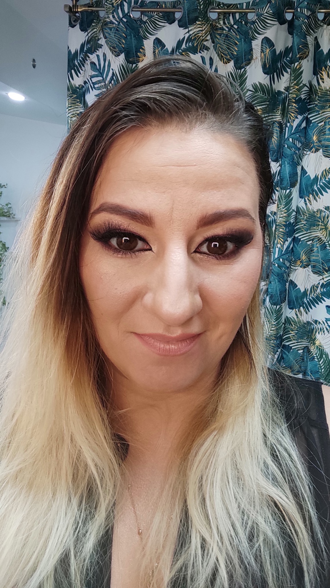 Portret kobiety z pełnym makijażem oczu w odcieniach brązu i długimi sztucznymi rzęsami, na tle zasłony w tropikalne liście.