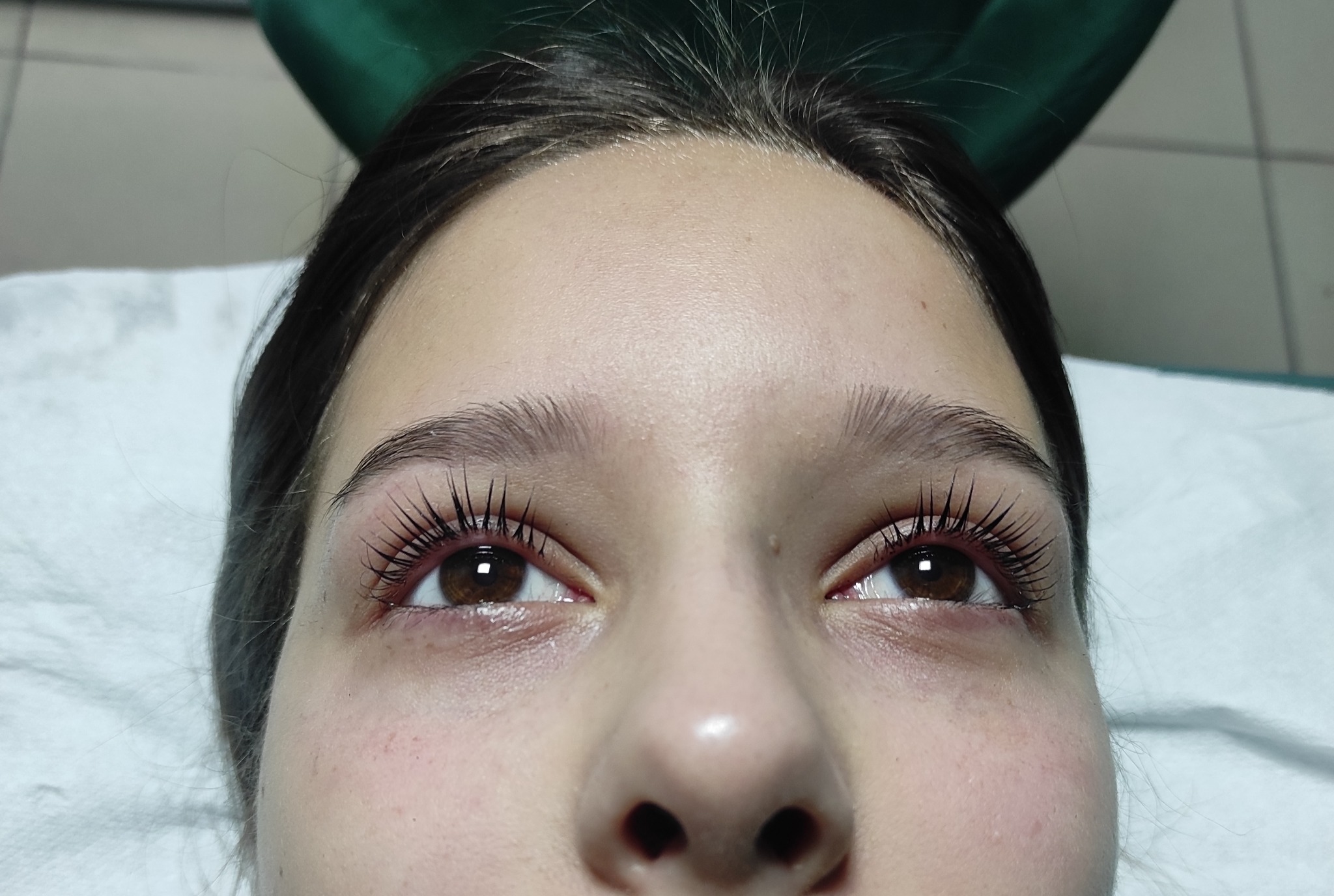 Zbliżenie na twarz kobiety po zabiegu liftingu rzęs, widoczne wydłużone i podkręcone rzęsy, brązowe oczy, skóra z delikatnymi zaczerwienieniami w okolicy oczu, ujęcie z dołu.