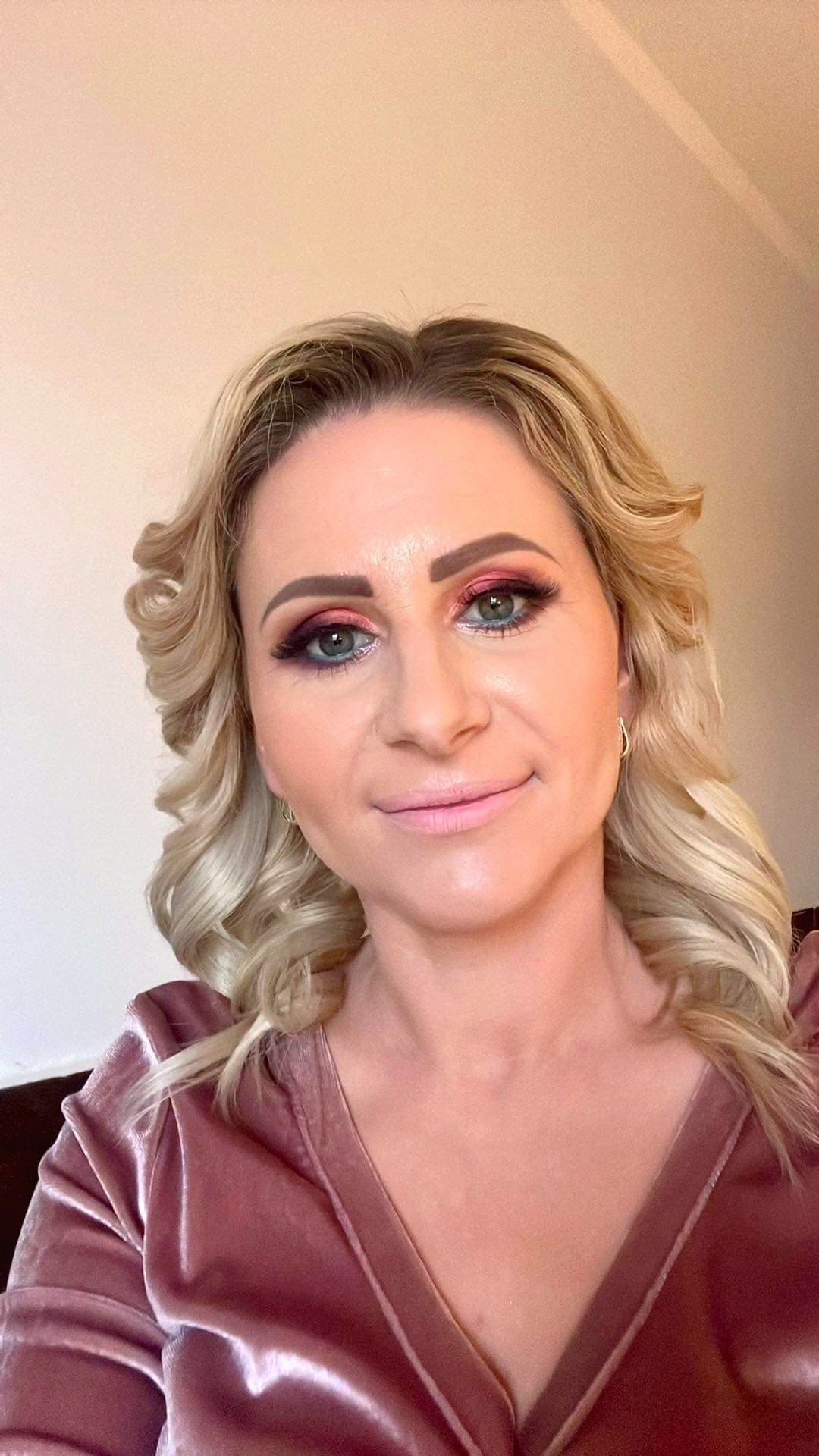 Portret kobiety z profesjonalnym makijażem oczu w odcieniach różu i fioletu, podkreślonymi rzęsami i delikatnie zaznaczonymi ustami w błyszczącym różowym topie z weluru. Blond włosy ułożone w loki.