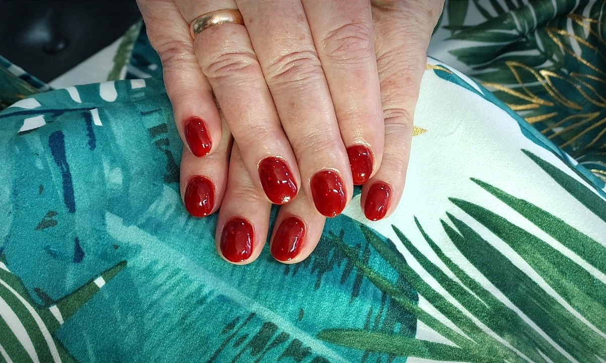 Dłonie z czerwonym, błyszczącym manicure hybrydowym na tle tkaniny z motywem tropikalnych liści; widoczny złoty pierścionek na palcu serdecznym.