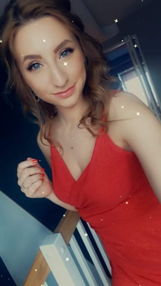 Portret kobiety w czerwonej sukience z subtelnym makijażem oczu i pomalowanymi na czerwono paznokciami, stojącej przy białych schodach.