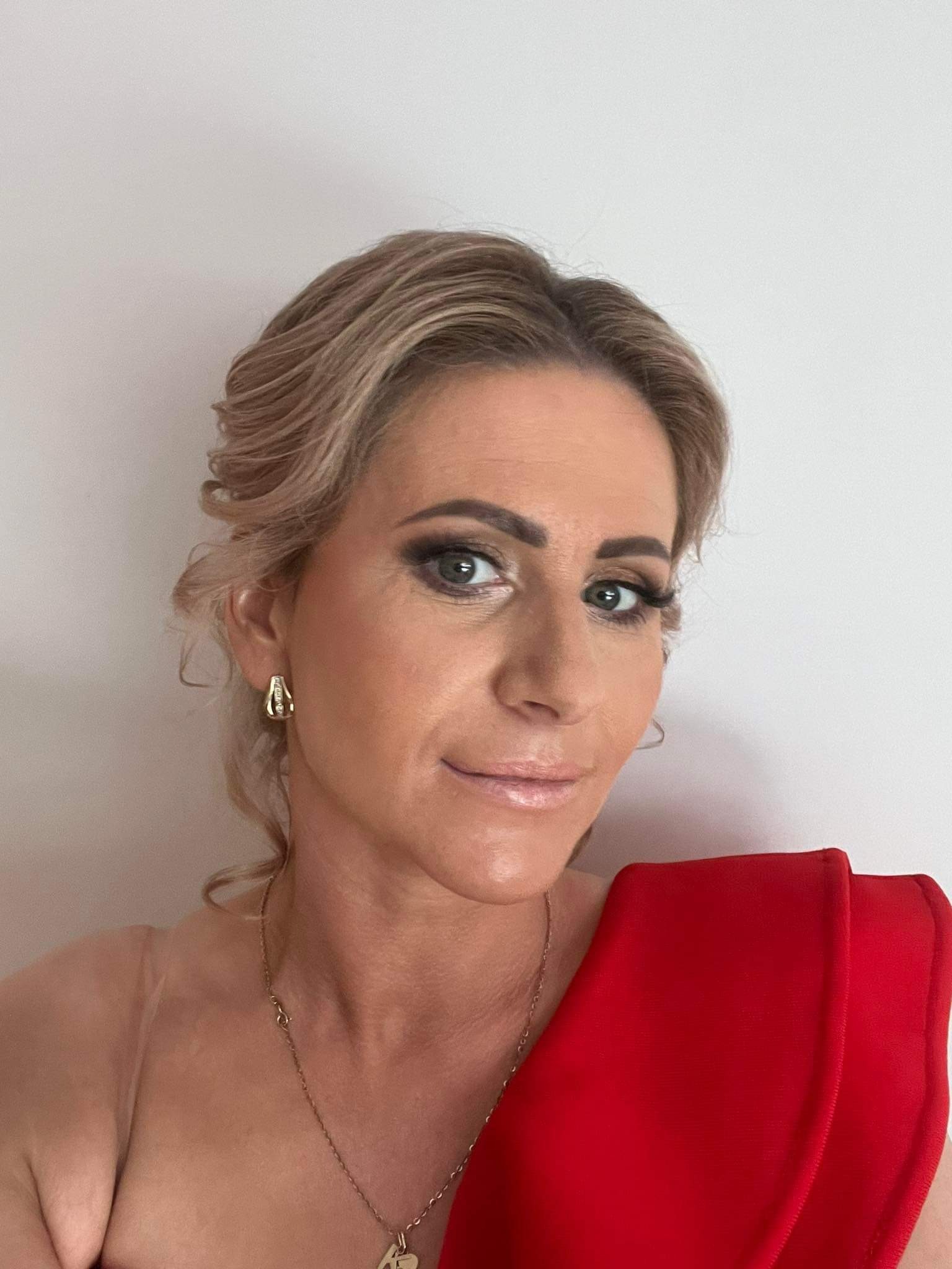 Portret kobiety z profesjonalnym makijażem, blond włosami upiętymi do góry, złotą biżuterią i czerwoną sukienką na białym tle.