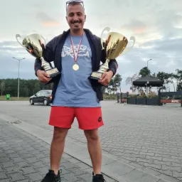 Uśmiechnięty mężczyzna w sportowym stroju, z okularami na głowie i medalem na szyi, trzymający dwa złote puchary z tabliczkami 'II MIEJSCE', na tle parkingu z zaparkowanym samochodem i budynkiem KFC.