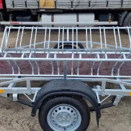 Przyczepka rowerowa z metalową konstrukcją do transportu rowerów, widoczna na tle czerwonej ciężarówki i ziemistego podłoża.