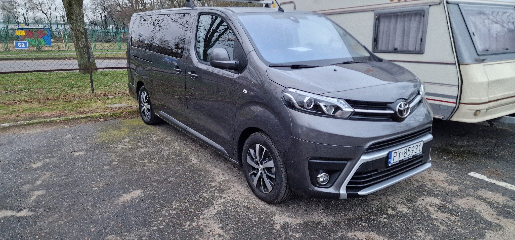Szary minivan Toyota Proace Verso zaparkowany obok przyczepy kempingowej na nierównym, asfaltowym terenie.