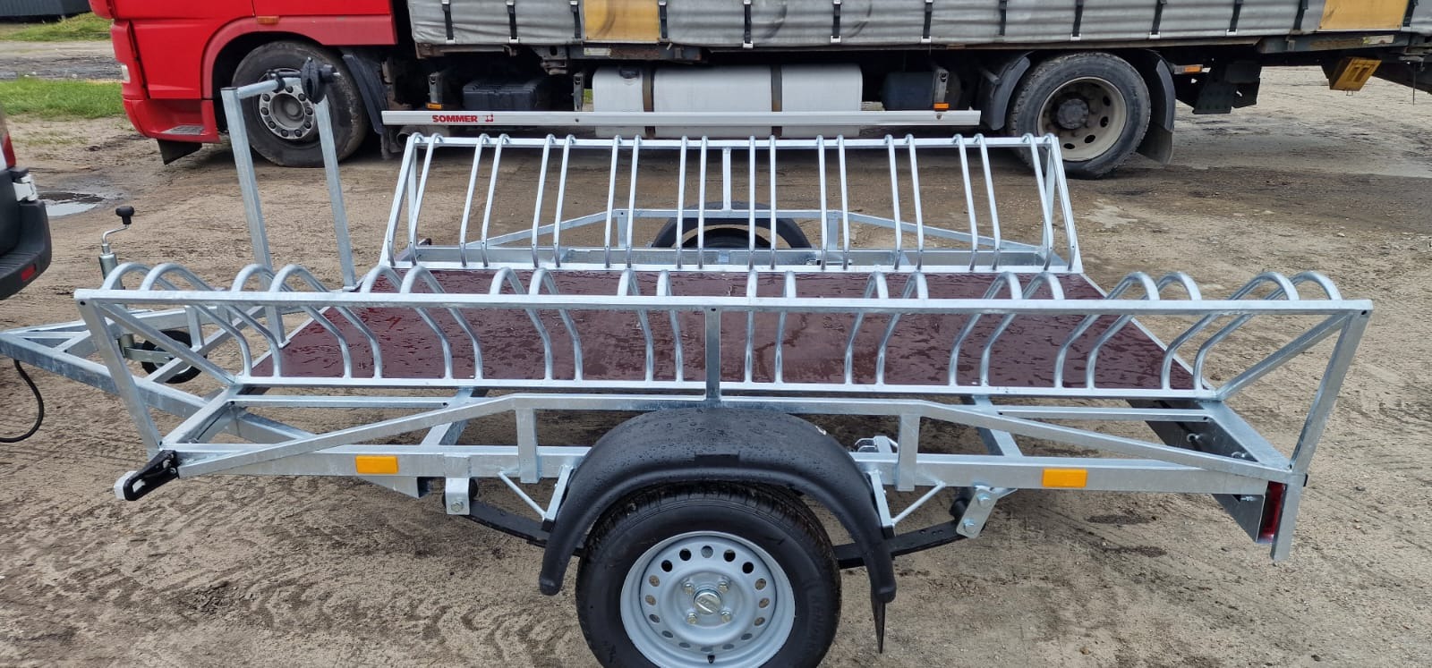 Przyczepka rowerowa z metalową konstrukcją do transportu rowerów, widoczna na tle czerwonej ciężarówki i ziemistego podłoża.