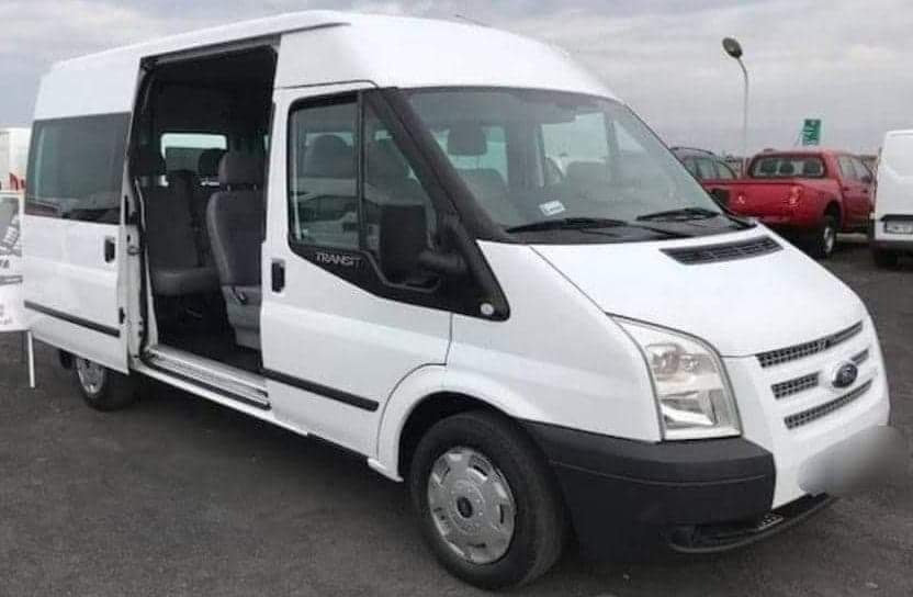 Biały bus marki Ford Transit z otwartymi przesuwnymi drzwiami bocznymi, widoczne szare fotele wewnątrz.