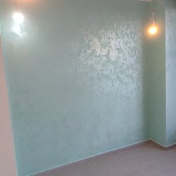 Serwis remontowy BEZ-VAS - Ściana w pokoju po malowaniu dekoracyjnym w odcieniach zieleni, z widoczną fakturą i oświetleniem punktowym. Szara podłoga i fragment instalacji przy ścianie.