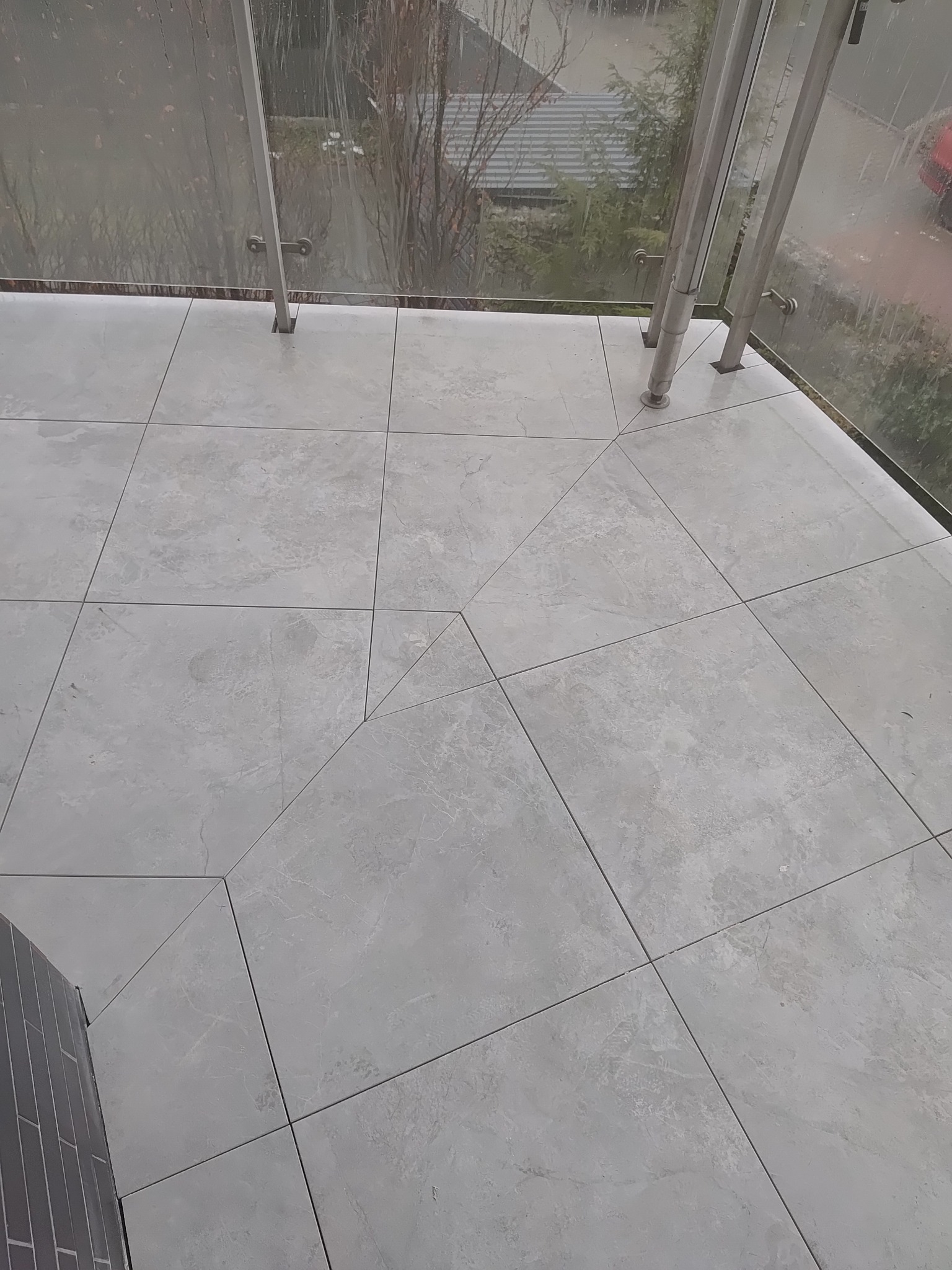 Jasnoszare płytki gresowe na balkonie z szklaną balustradą. Widok z góry, ukazujący wzór ułożenia płytek i minimalistyczny design. Nowoczesne wykończenie.