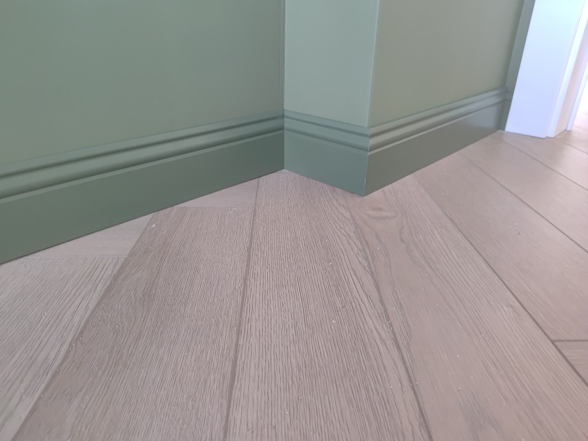 Ściana w odcieniach zieleni z listwami przypodłogowymi i panelami podłogowymi w kolorze jasnego drewna. Widoczny narożnik ściany i fragment białych drzwi.