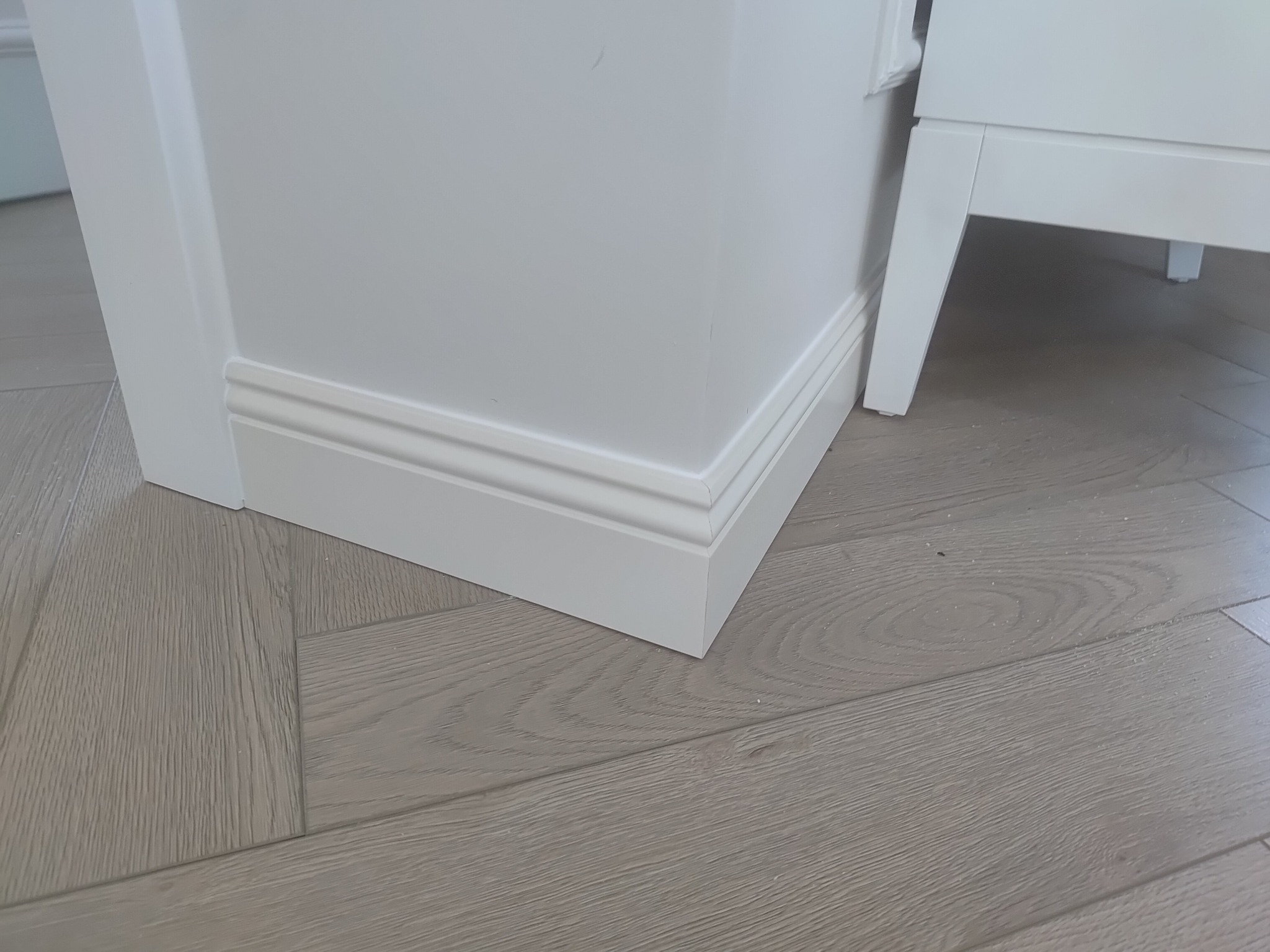 Biała listwa przypodłogowa z frezami, zamontowana przy ścianie i drzwiach, na podłodze z paneli w odcieniach szarości. Fragment mebla w tle.