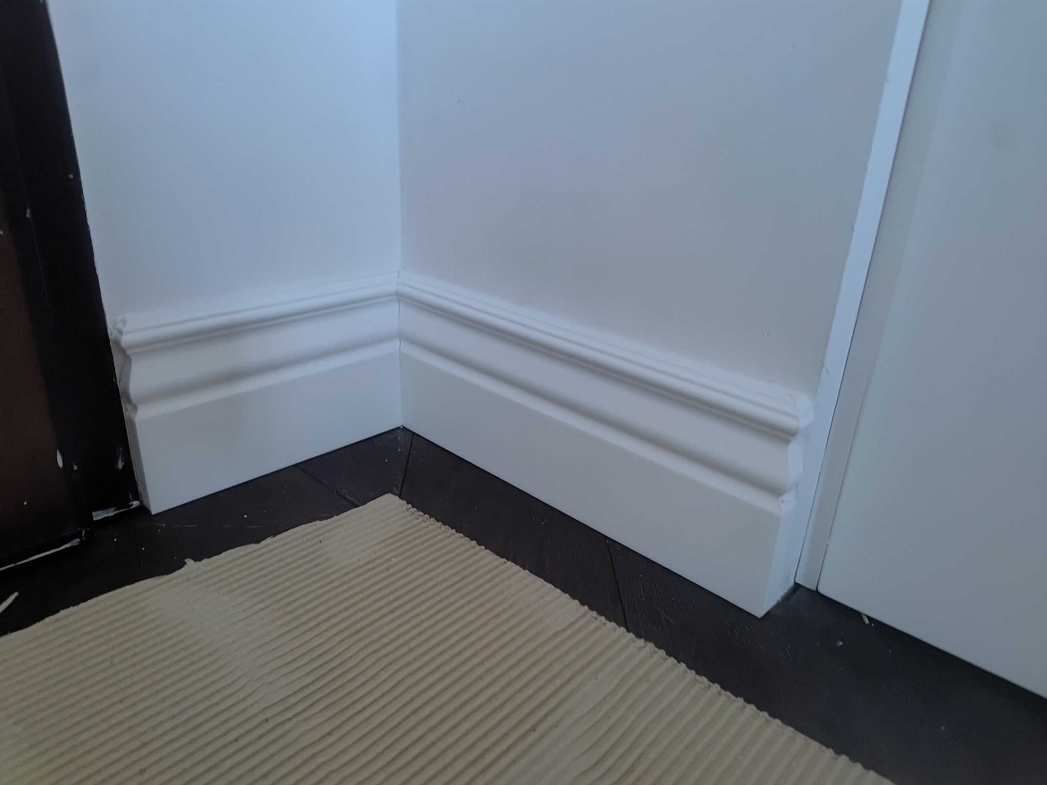 Białe listwy przypodłogowe zamontowane w rogu pokoju, widoczne na tle ciemnej podłogi i fragmentu zabezpieczonej powierzchni. Detal wykończenia wnętrza.