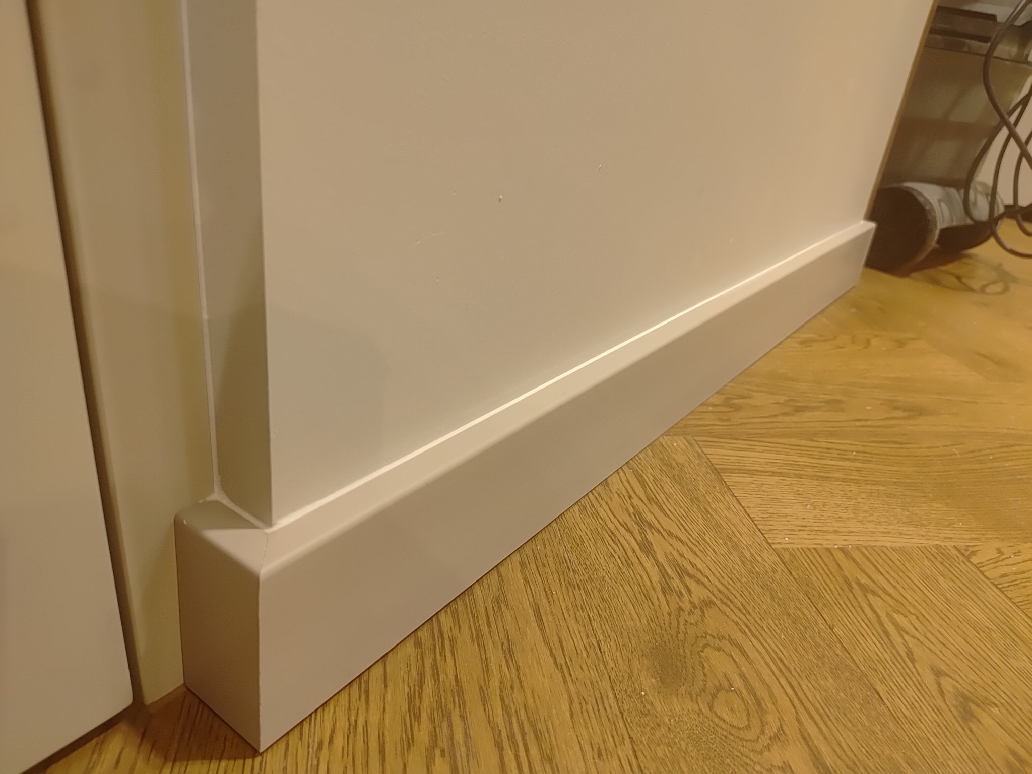 Zamontowana, pomalowana na jasny kolor listwa przypodłogowa przy ścianie i drewnianej podłodze. Widoczny fragment drzwi po lewej stronie.