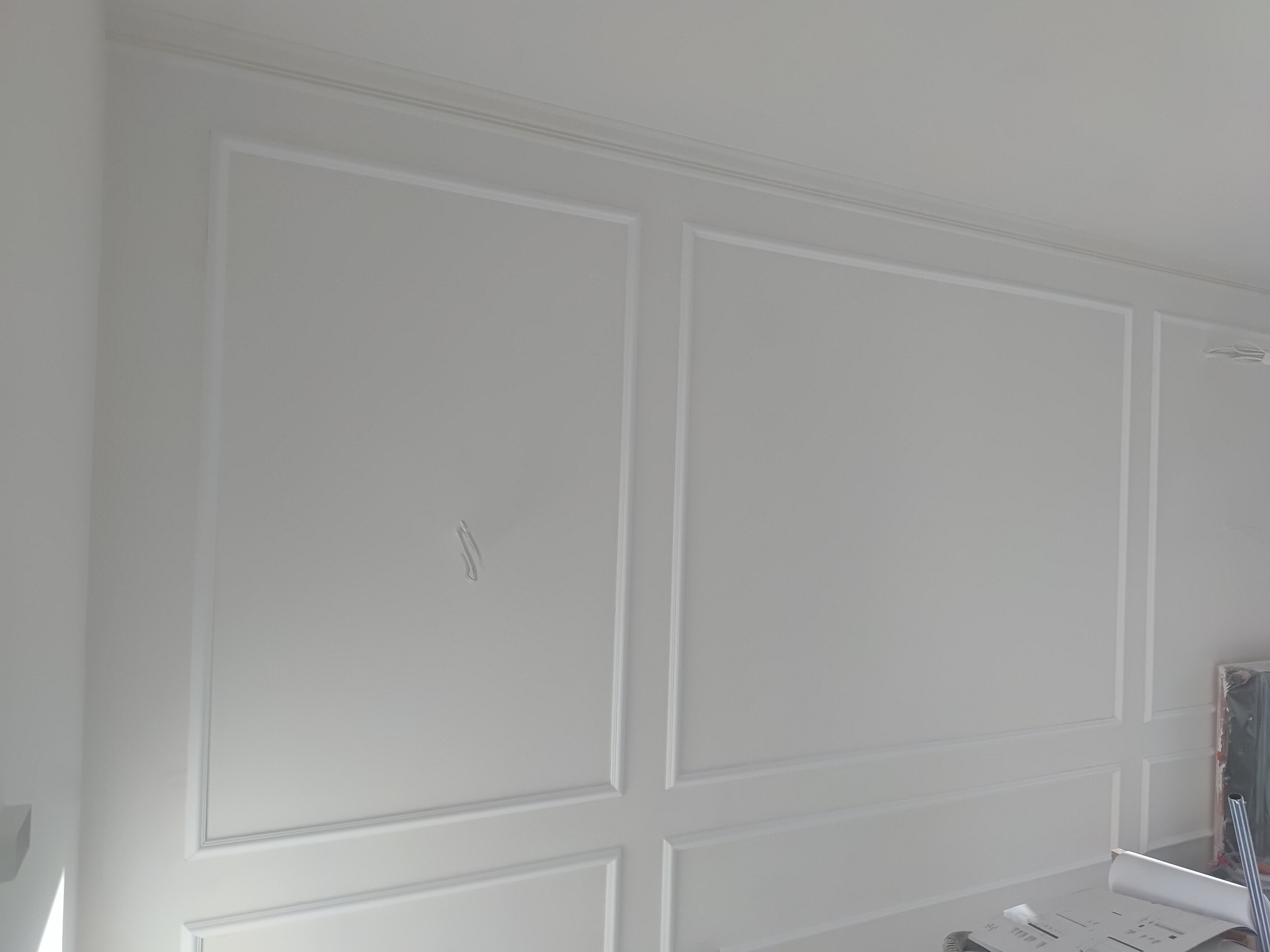 Biała ściana z dekoracyjnymi panelami i listwami, tworzącymi geometryczne wzory. Minimalistyczny design, świeżo po montażu, z widocznymi detalami wykończenia.