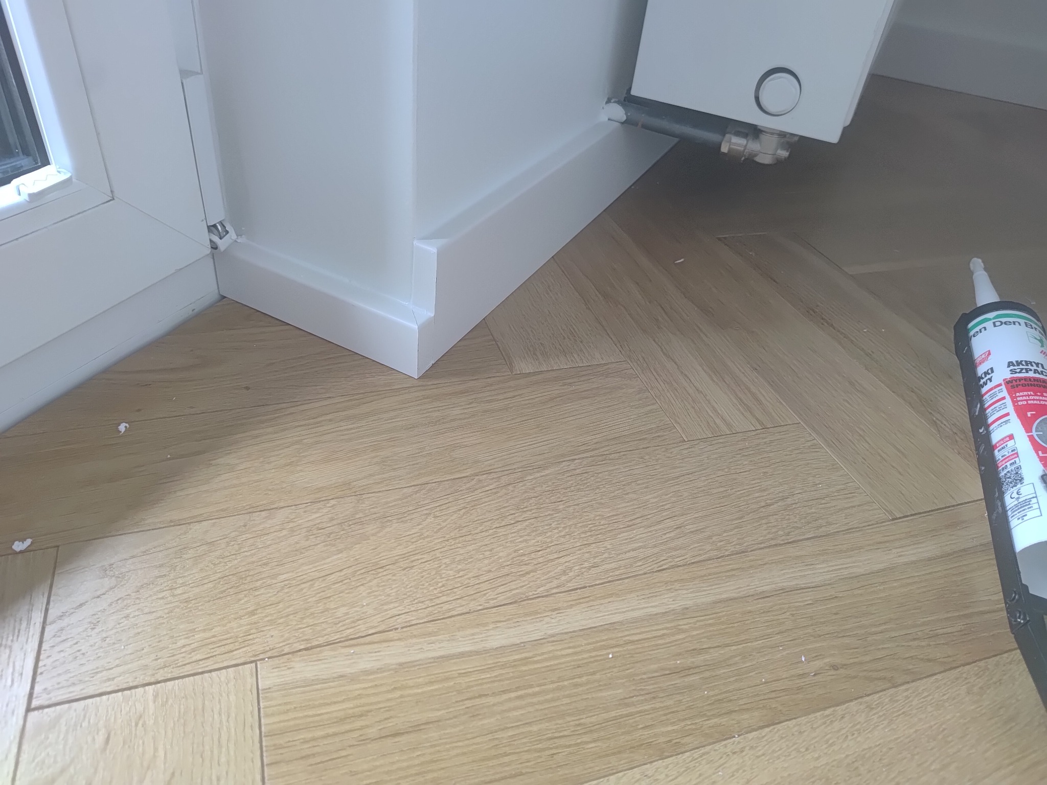 Ukończone białe listwy przypodłogowe przy oknie i ścianie na parkiecie ułożonym w jodełkę. Widoczny akryl szpachlowy.