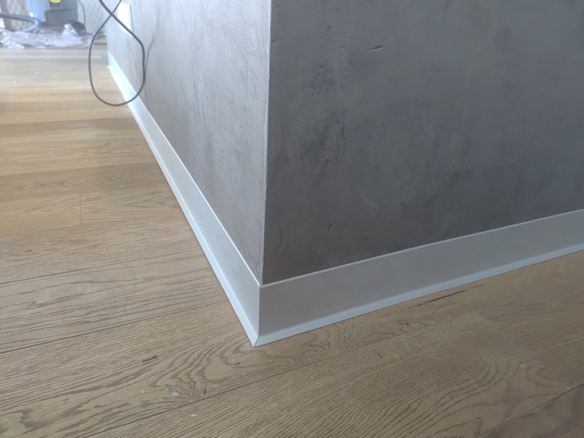 Wykończona ściana z listwą przypodłogową w Piasecznie. Szara ściana o fakturze betonu, drewniana podłoga. Detal wykończenia wnętrza, estetyczne połączenie powierzchni.