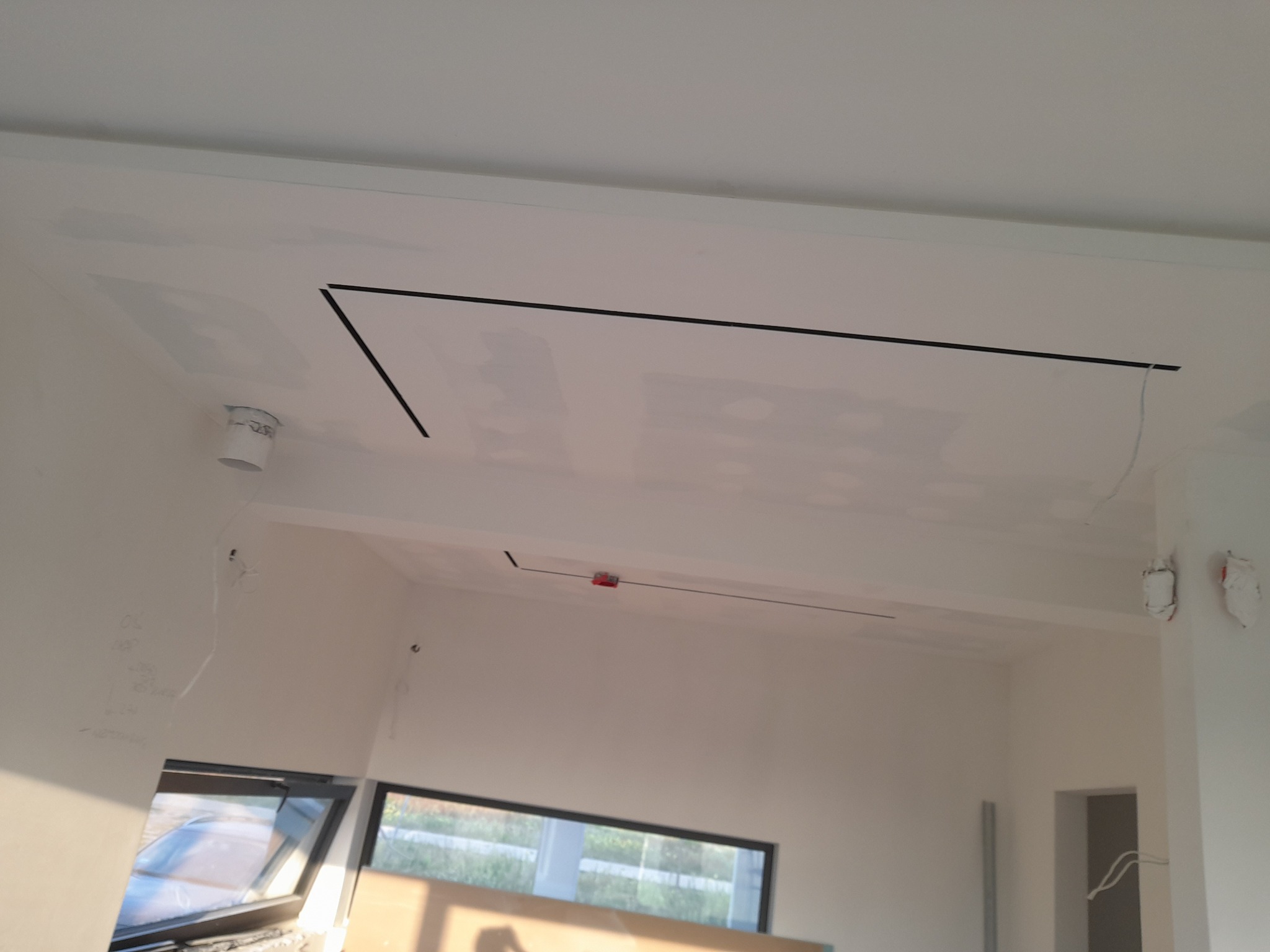 Biały sufit z płyt kartonowo-gipsowych, widoczne czarne linie wyznaczające kształt, okno w tle. Wnętrze w trakcie wykończenia.