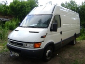 Biały samochód dostawczy Iveco z widocznymi tablicami rejestracyjnymi, zaparkowany na trawiastym terenie, prawdopodobnie przed budynkiem lub w lesie, sugerujący usługi transportowe...