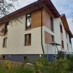 Dwukondygnacyjny dom z elewacją w kolorze kremowym i panelami imitującymi drewno, widoczne rusztowanie przy ścianie bocznej, ciemna podmurówka i okna z drewnianymi ramami.