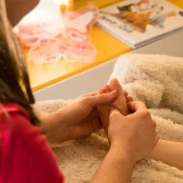 Terapeuta wykonuje delikatny masaż stóp dziecka na miękkim, kremowym kocu; w tle widoczny żółty stolik z dziecięcymi dekoracjami i książką.