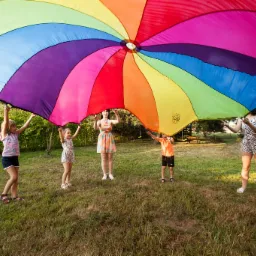 Grupa dzieci i animatorka trzymająca kolorową chustę animacyjną na trawniku podczas zabawy na świeżym powietrzu.