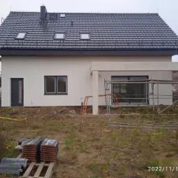 Nowo wybudowany dom jednorodzinny z szarą dachówką i białymi ścianami, widoczne rusztowania i paleta z dachówkami na pierwszym planie.