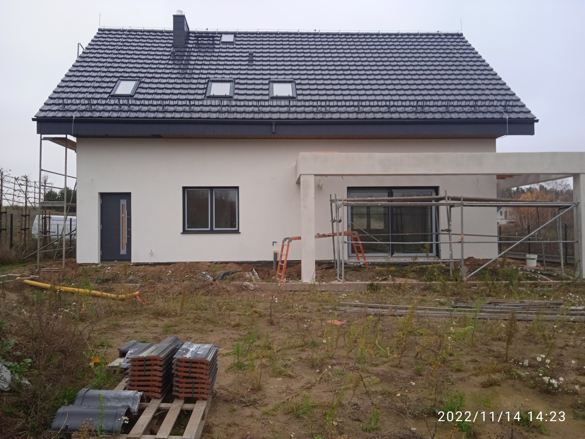Nowo wybudowany dom jednorodzinny z szarą dachówką i białymi ścianami, widoczne rusztowania i paleta z dachówkami na pierwszym planie.