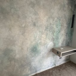 Elmat firma usługowo handlowa - Ściana z dekoracyjną, nieregularną fakturą w odcieniach szarości i zieleni, widoczna półka z żarówką i fragment podłogi z brązowymi panelami.